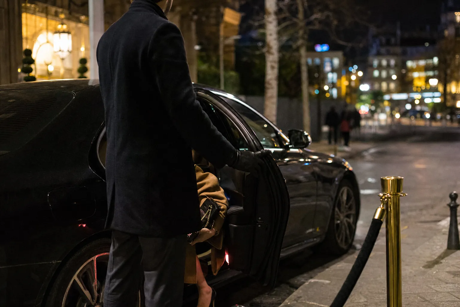 plan large d'une berline noire garee le long d'un trottoir devant l'entree eclairee d'un hotel de standing la nuit, portiere